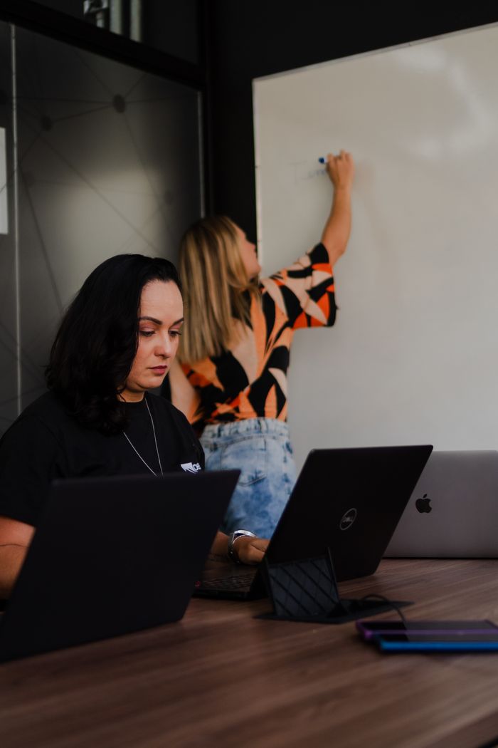 Two women engaged in a collaborative office workspace, one writing on a whiteboard.